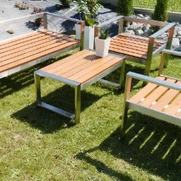 Zestaw mebli ogrodowych: ławka, dwa krzesła i stolik kawowy, wykonane z drewna i stali nierdzewnej, ustawione na trawie w ogrodzie, z roślinami w donicach w tle.