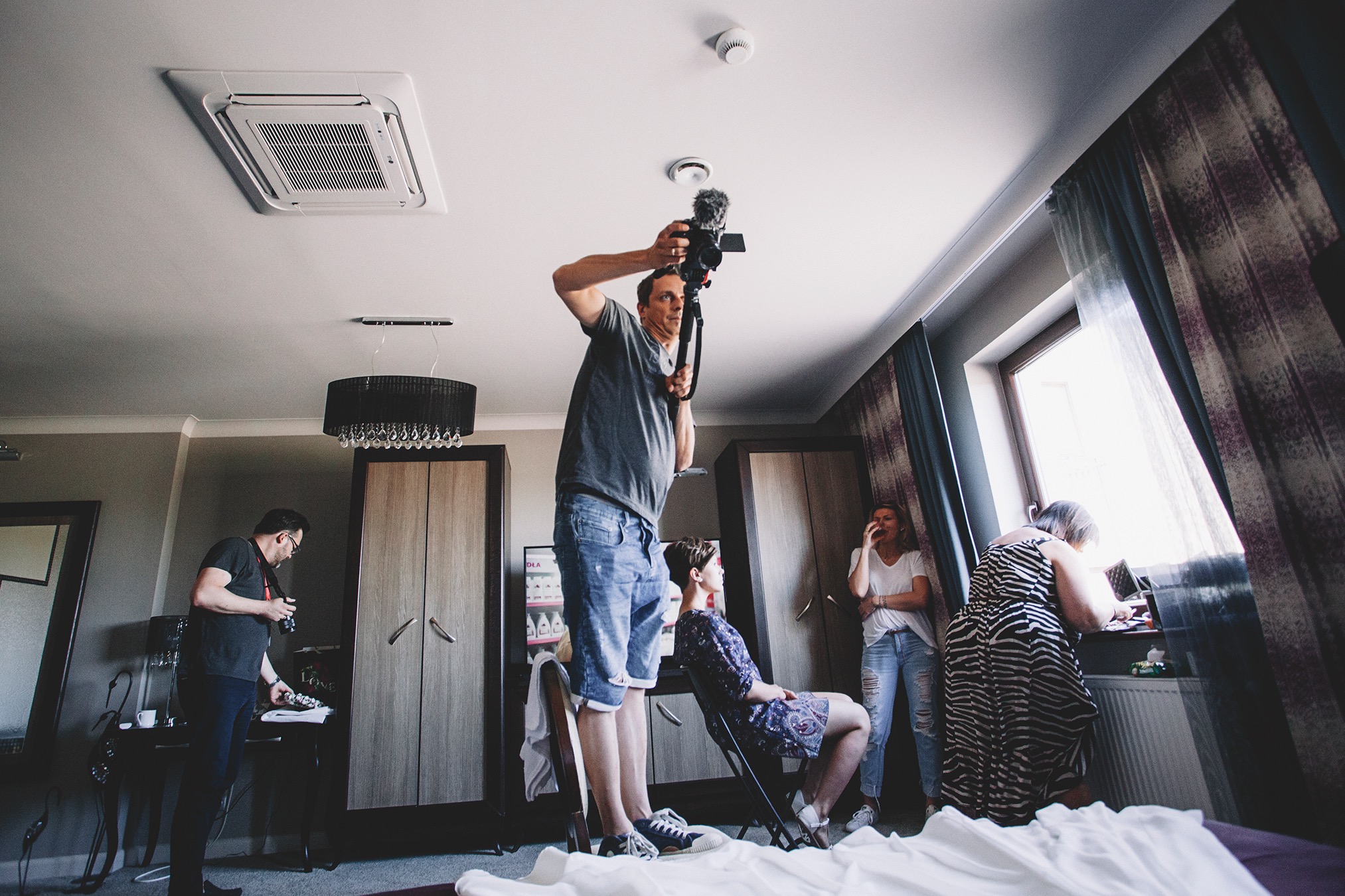 Ekipa filmowa w pokoju hotelowym podczas kręcenia ujęć z udziałem kilku osób, oświetlenie dzienne wpada przez okno.