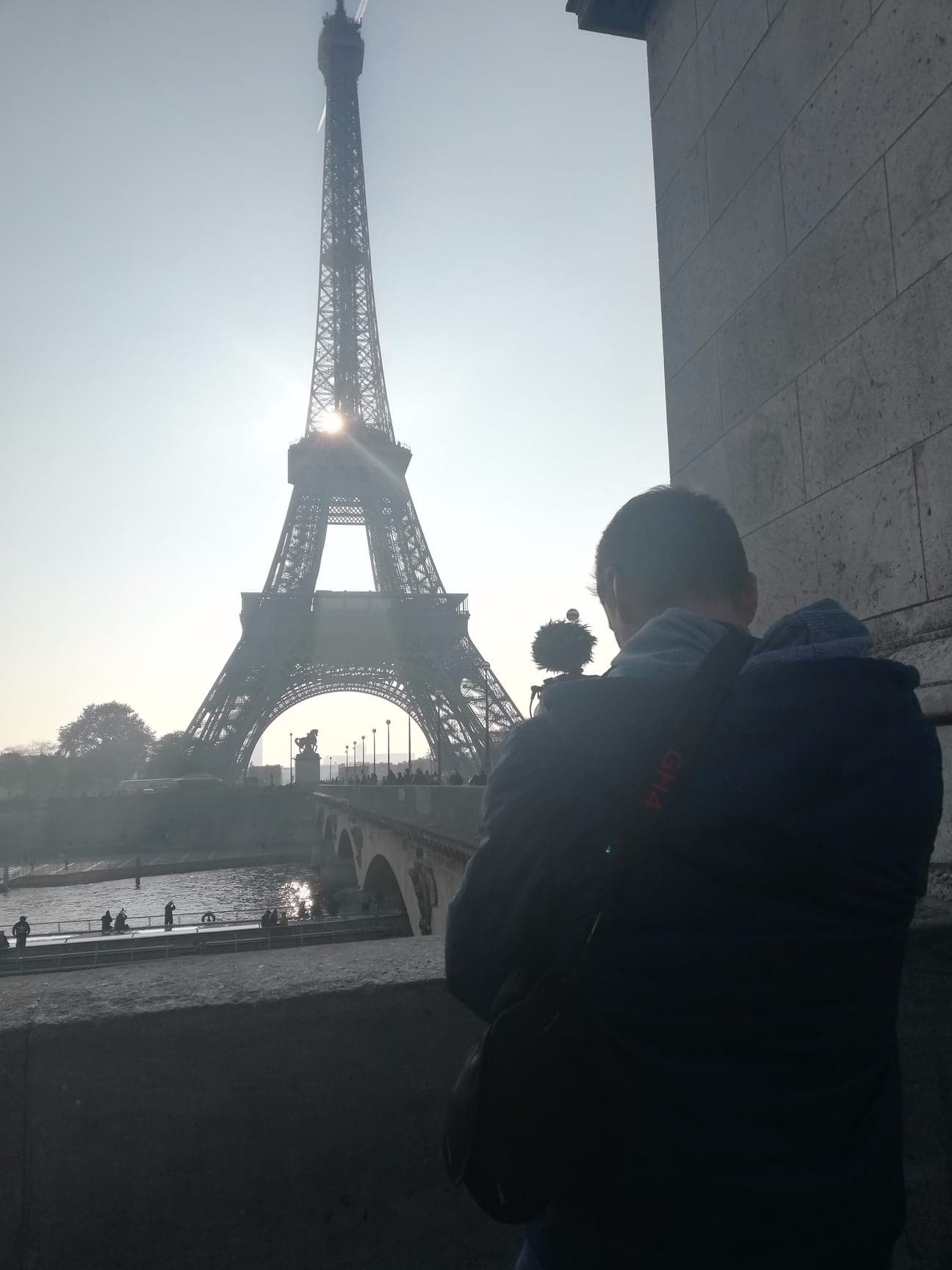 Osoba z mikrofonem kierunkowym na ramieniu, stojąca na tle wieży Eiffla i mostu w Paryżu, podczas słonecznego dnia.