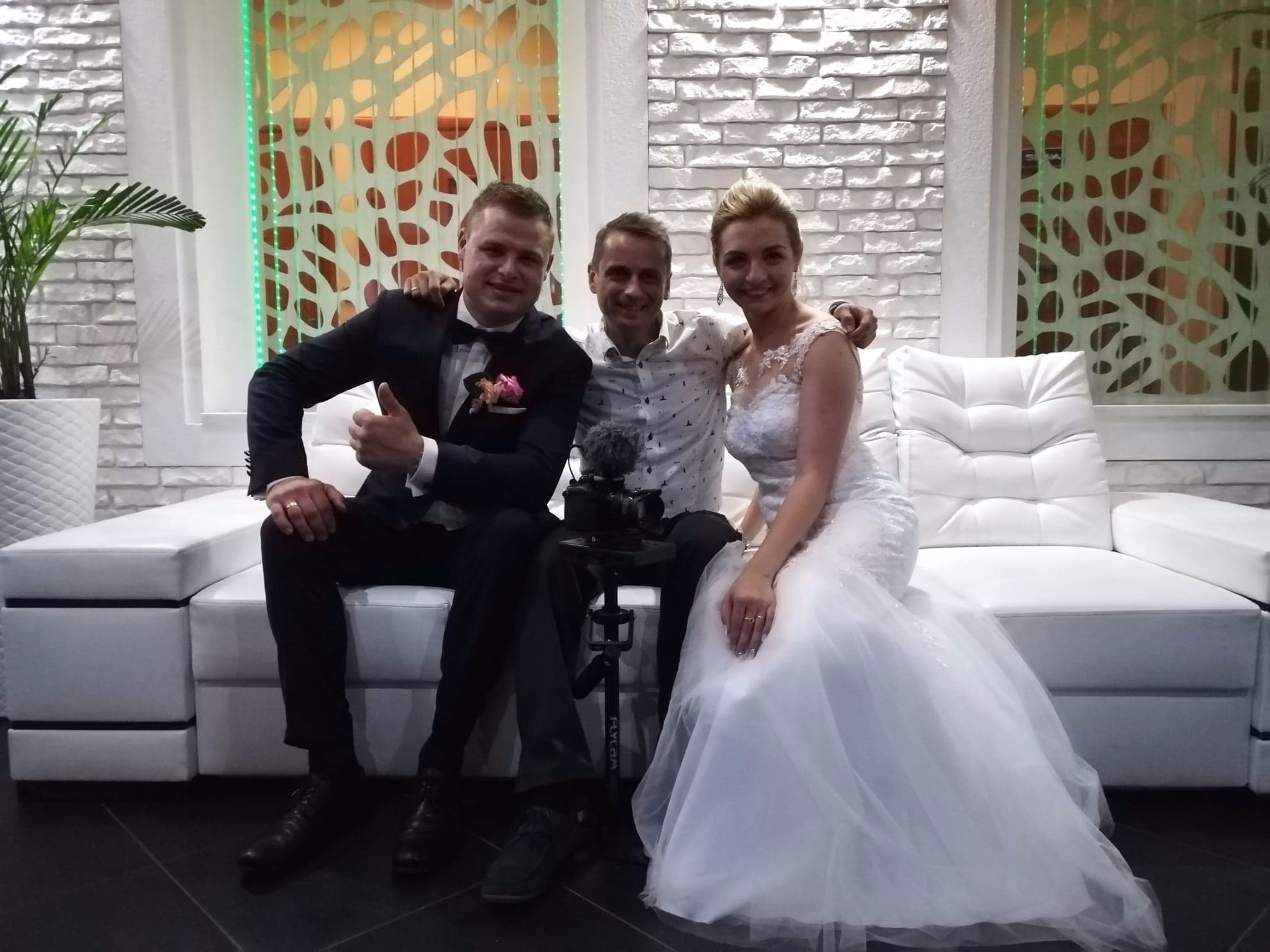 Uśmiechnięta para młoda w sukni ślubnej i garniturze pozuje z osobą z mikrofonem i kamerą na statywie siedząc na białej sofie przed ścianą z białej cegły i dekoracyjnymi panelami.