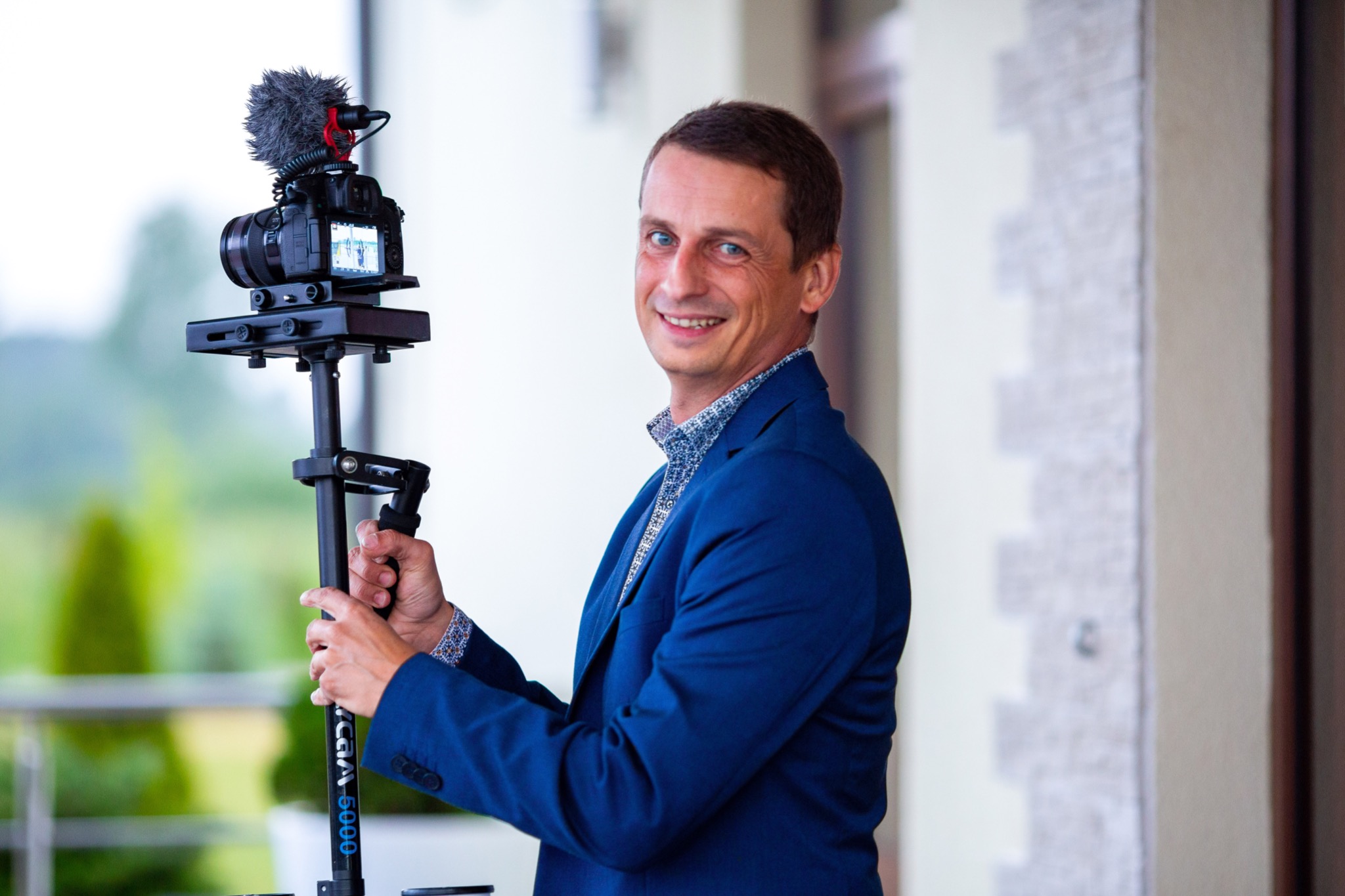 Uśmiechnięty kamerzysta w niebieskiej marynarce, trzymający stabilizator z zamontowaną kamerą z mikrofonem typu 'deadcat', na tle jasnej ściany i zieleni.