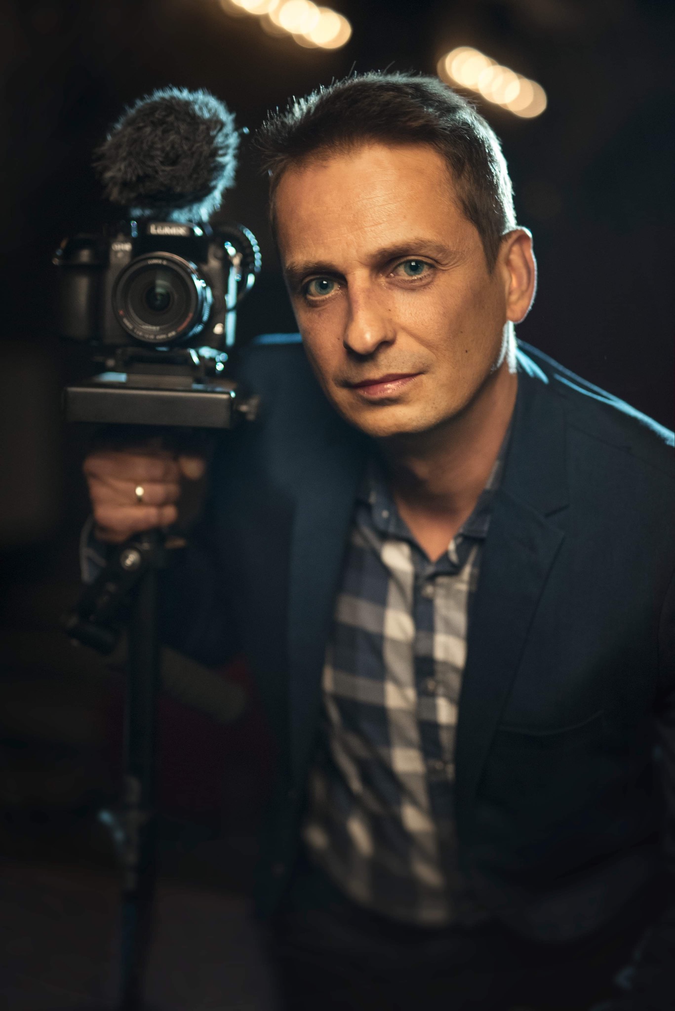 Portret mężczyzny z kamerą Lumix i mikrofonem Rode na statywie, ubrany w niebieską marynarkę i koszulę w kratę, z pierścieniem na palcu.