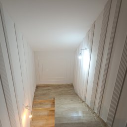 Usługi Remontowe Paweł Furgała - Nowoczesny korytarz z białymi, pionowymi panelami ściennymi o ryflowanej fakturze, jasną podłogą z beżowych płytek imitujących kamień i tymczasowym oświetleniem wiszącymi żarówkami.
