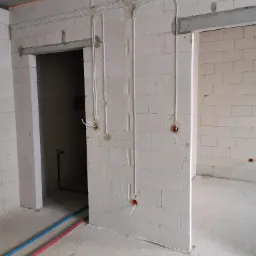 Wewnętrzne instalacje elektryczne w surowym stanie: białe rury prowadzone po ścianach z bloczków, widoczne kable wystające z puszek instalacyjnych, rury instalacji wodnej na podłodze, surowy...