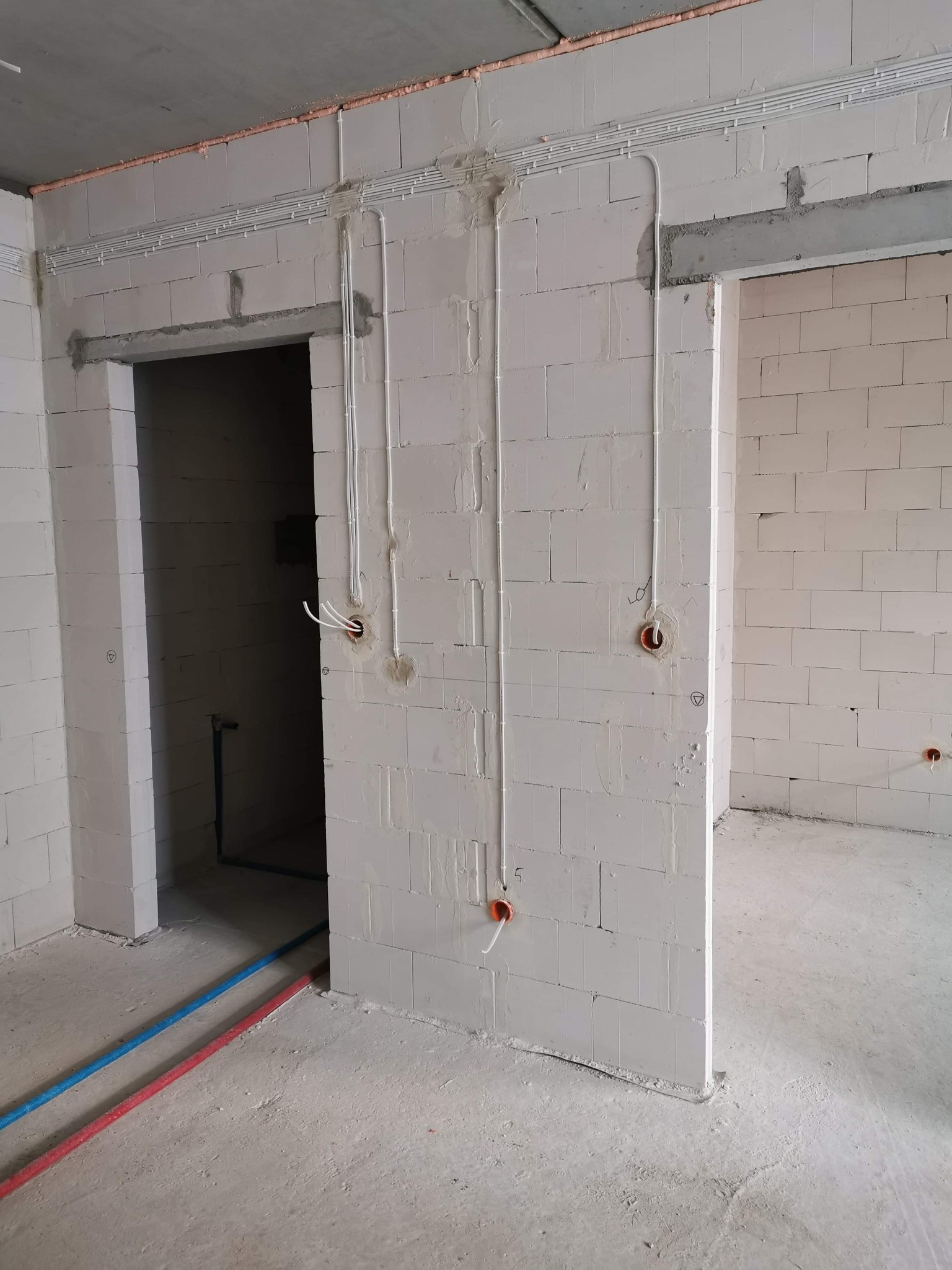 Wewnętrzne instalacje elektryczne w surowym stanie: białe rury prowadzone po ścianach z bloczków, widoczne kable wystające z puszek instalacyjnych, rury instalacji wodnej na podłodze, surowy...
