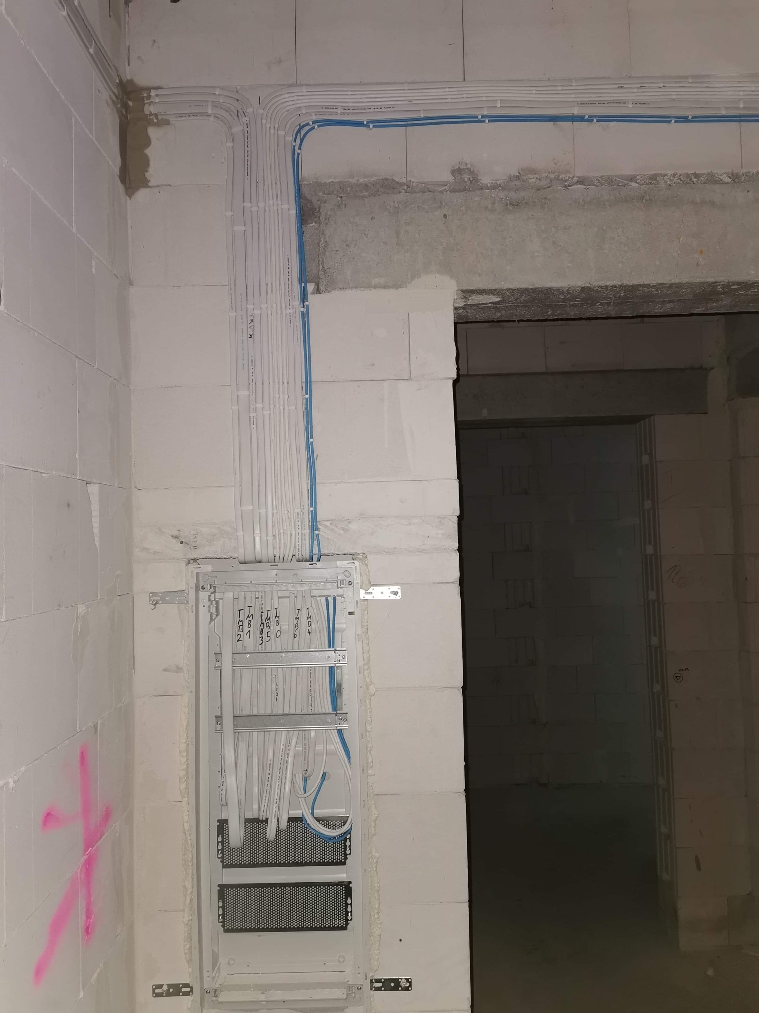 Rozprowadzenie przewodów elektrycznych w białych peszlach i niebieskich rurkach, zamocowanych na ścianie z białych bloczków, widoczna metalowa skrzynka rozdzielcza z czarnymi panelami...