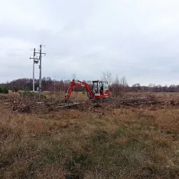 Czerwona mini koparka O&K w trakcie karczowania terenu z gałęzi i krzewów na zarośniętej łące, widoczny słup energetyczny w tle.