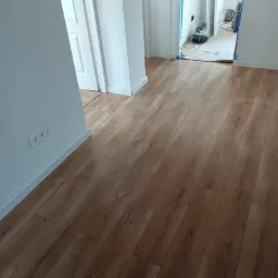 Ułożone panele podłogowe w jasnym pomieszczeniu z białymi ścianami i listwami przypodłogowymi, widoczne gniazdko elektryczne i otwarte drzwi z zabezpieczeniem folią malarską.