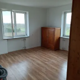 Pokój po odświeżeniu: świeżo pomalowane białe ściany, ułożone panele podłogowe, drewniana szafa, widoczne dwa okna z widokiem na zewnątrz i fragment odkurzacza w rogu.