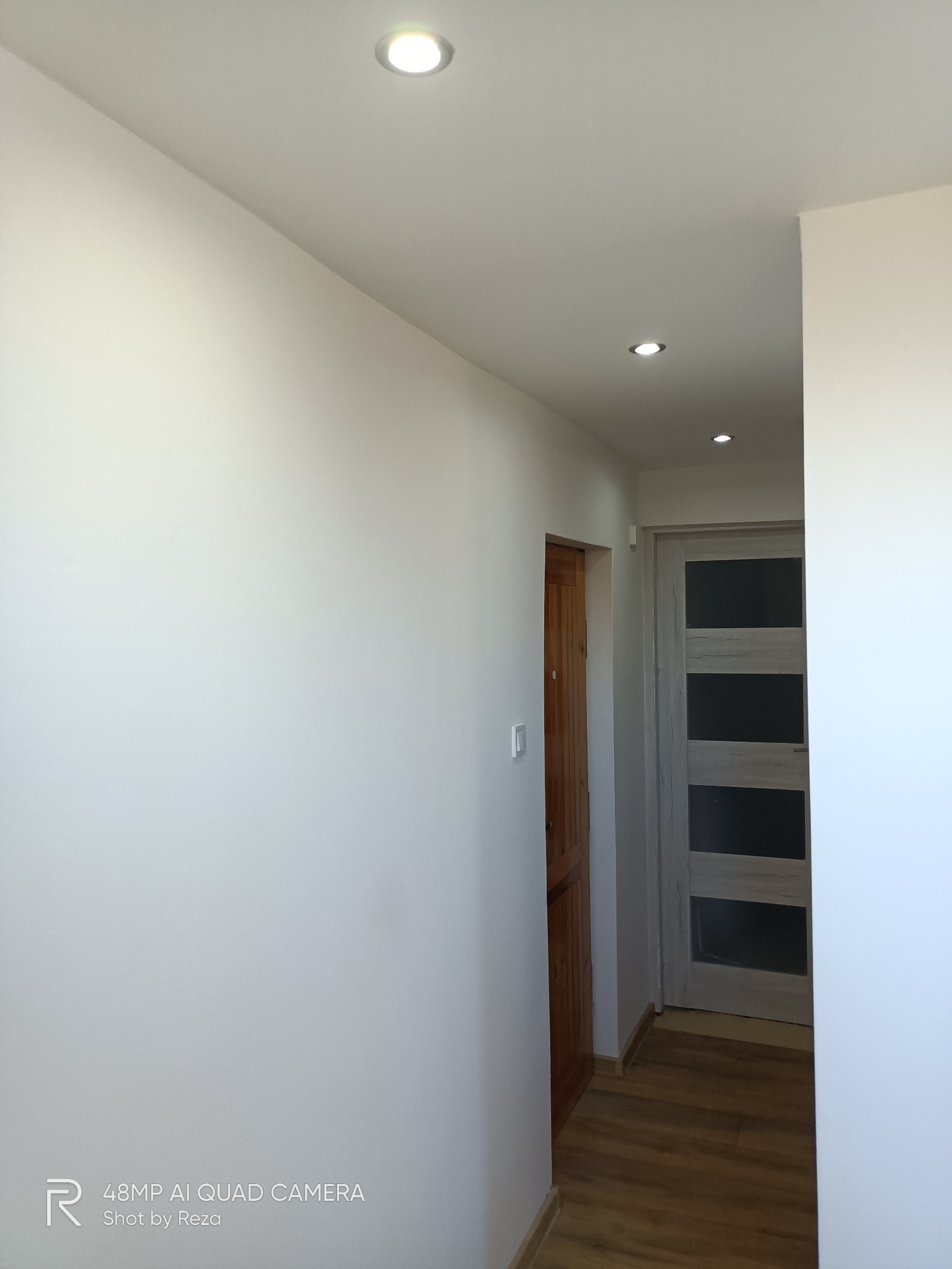 Korytarz po odświeżeniu: białe ściany, drewniana podłoga, dwoje drzwi - jedne drewniane, drugie z przeszkleniami, oświetlenie punktowe w suficie.