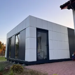 Gotowy modułowy budynek z białych i grafitowych płyt warstwowych, widoczne okna i drzwi z ciemną ramą, na zewnątrz butla z gazem i śmieci