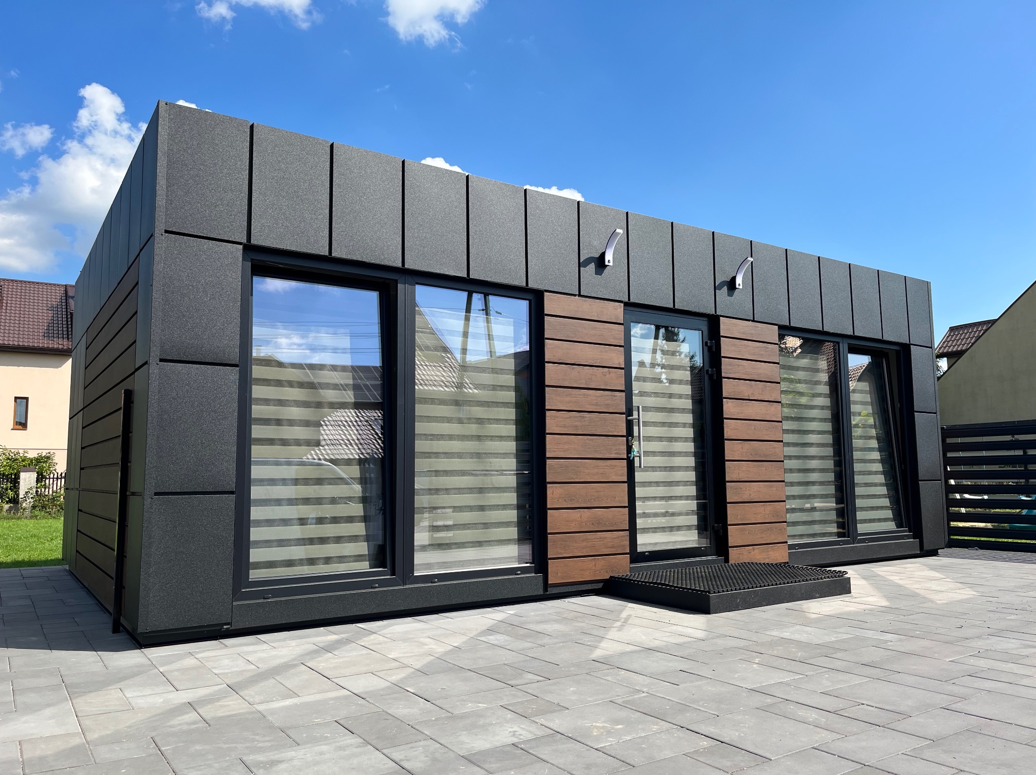 Nowoczesny, parterowy budynek modułowy z czarnych płyt warstwowych i drewnopodobnych paneli elewacyjnych, widoczne okna z żaluzjami poziomymi i kostka brukowa przed budynkiem w słoneczny dzień.
