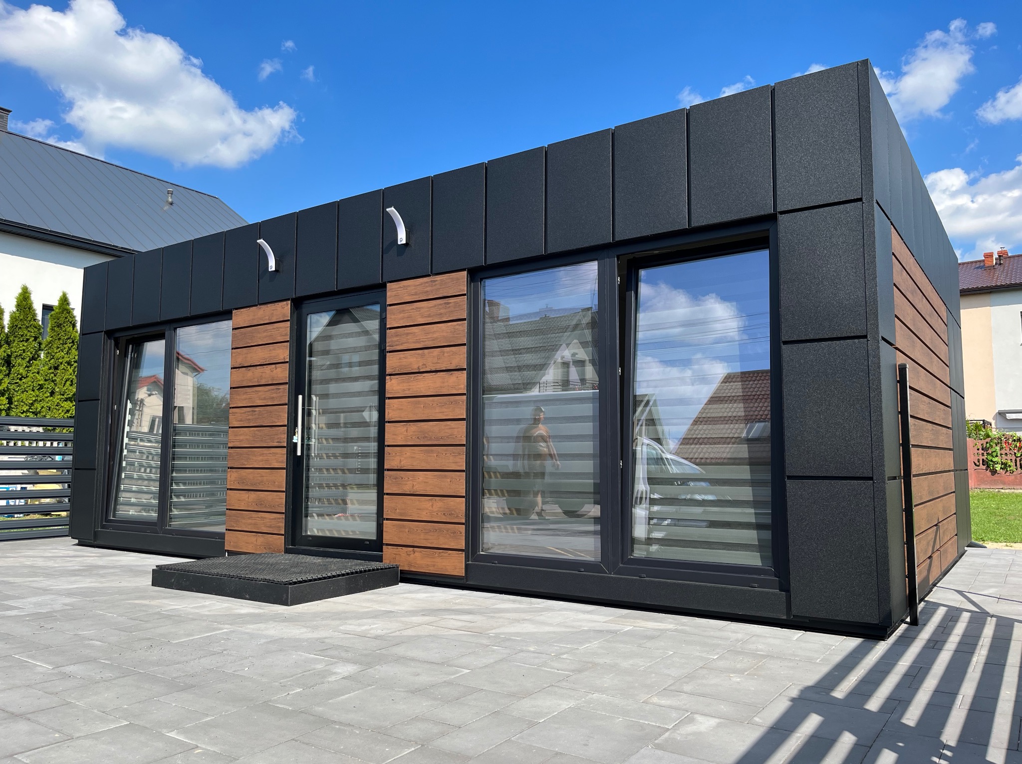 Nowoczesny, parterowy budynek modułowy z czarną elewacją z płyt warstwowych i drewnopodobnymi akcentami, duże okna z żaluzjami, kostka brukowa przed budynkiem, błękitne niebo z chmurami w tle.
