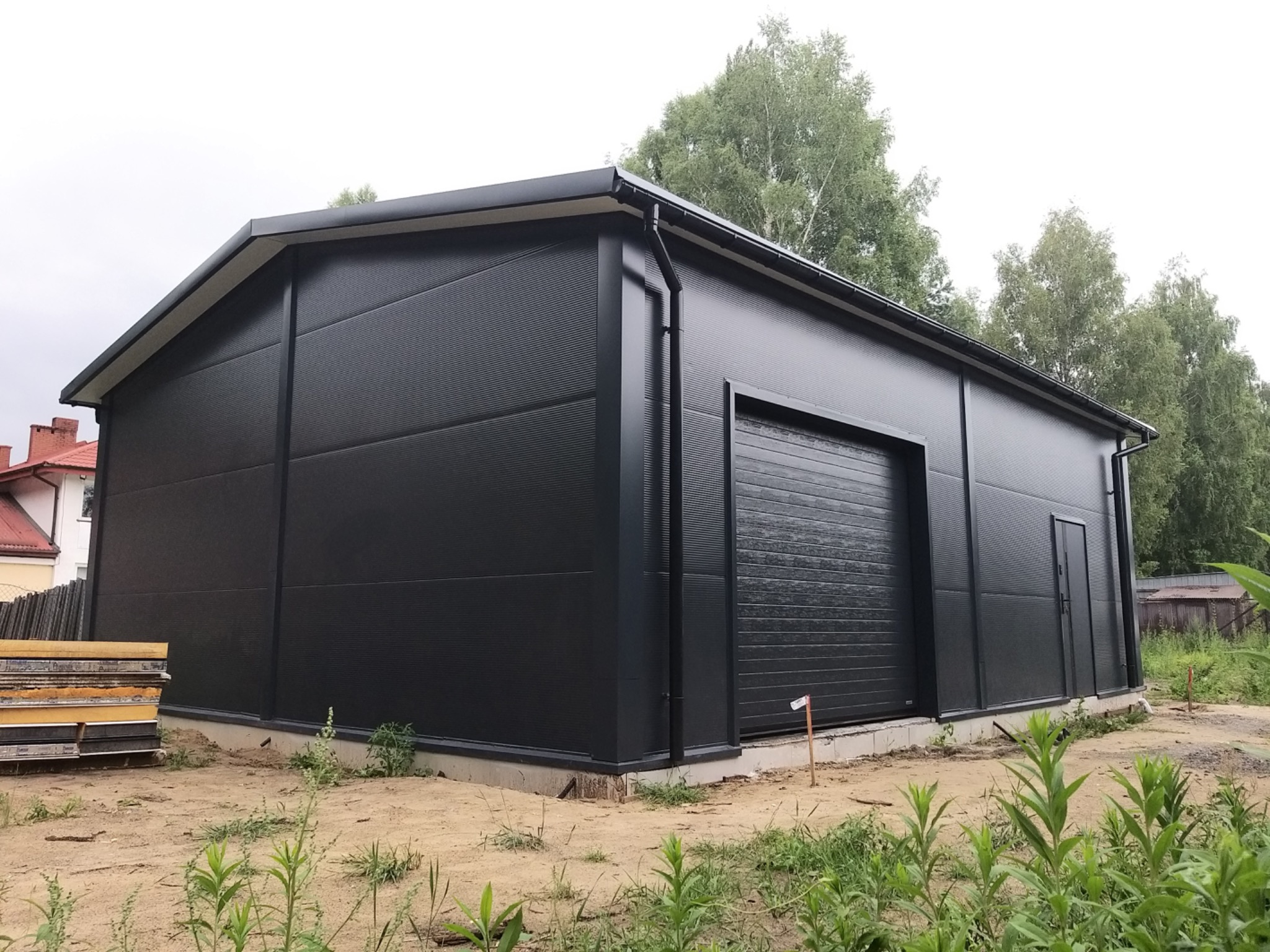 Czarny budynek przemysłowy z płyt warstwowych z bramą garażową i rynnami, widok zewnętrzny w pochmurny dzień.