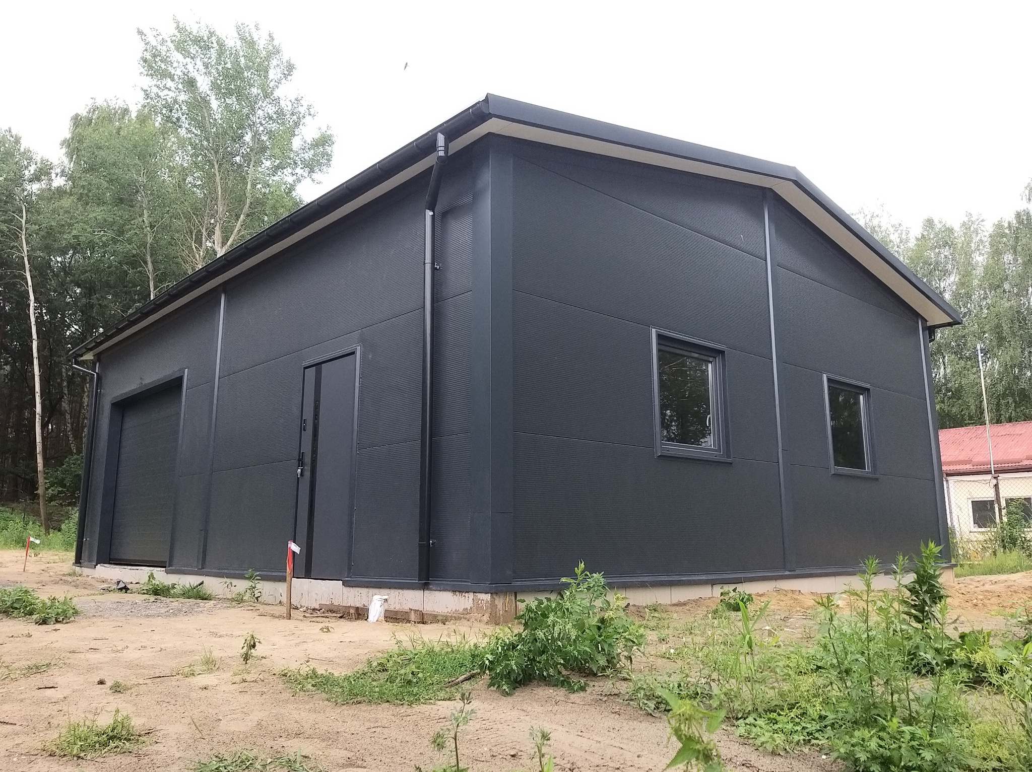 Gotowy budynek przemysłowy z ciemnoszarej płyty warstwowej z dwiema bramami garażowymi i dwoma oknami, widoczne obróbki blacharskie dachu i rynna spustowa, teren wokół budynku częściowo...