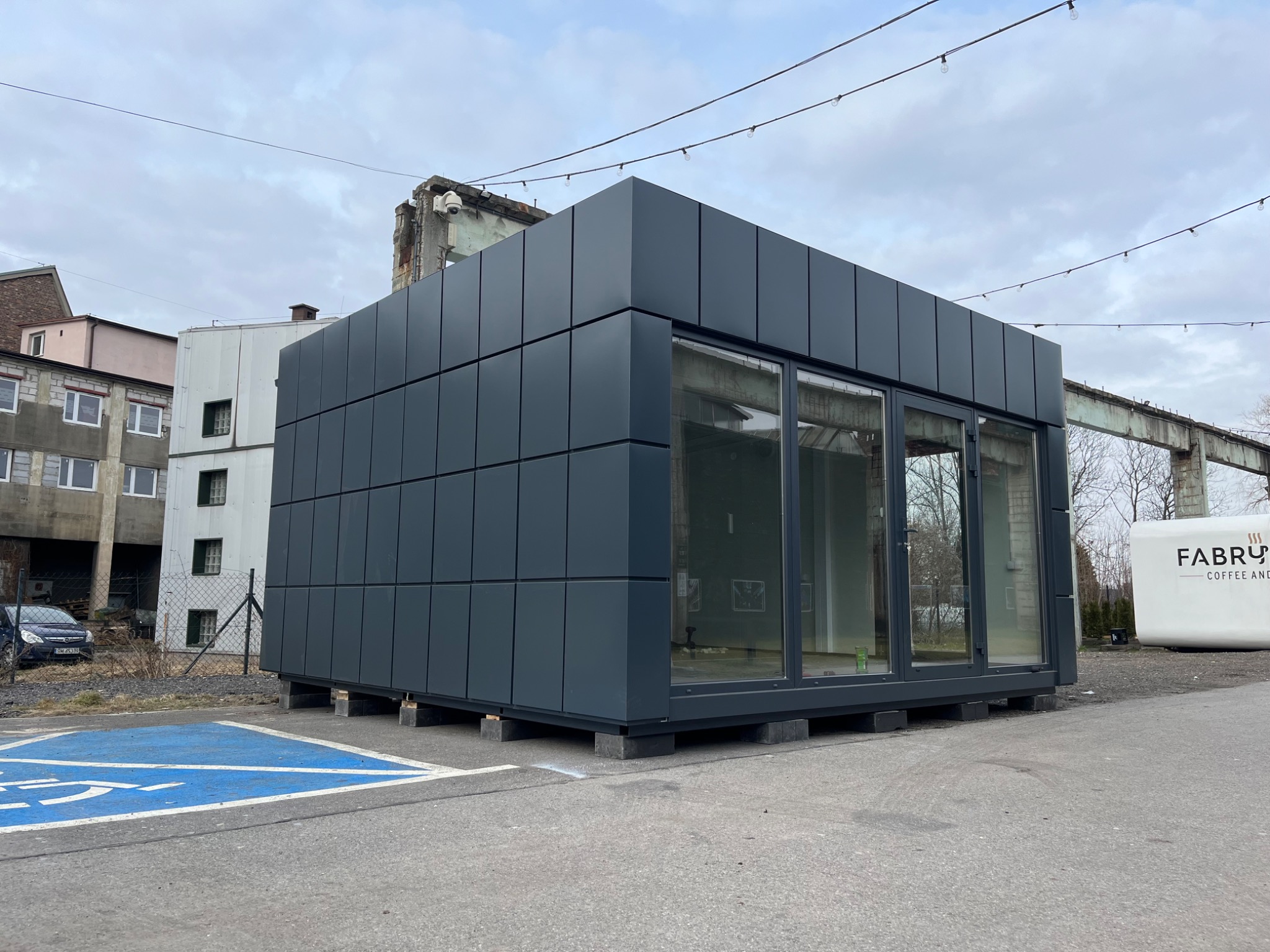 Nowoczesny pawilon handlowy z płyt warstwowych, ustawiony na betonowych bloczkach na parkingu z wyznaczonym miejscem dla niepełnosprawnych, w tle budynki i konstrukcja stalowa.