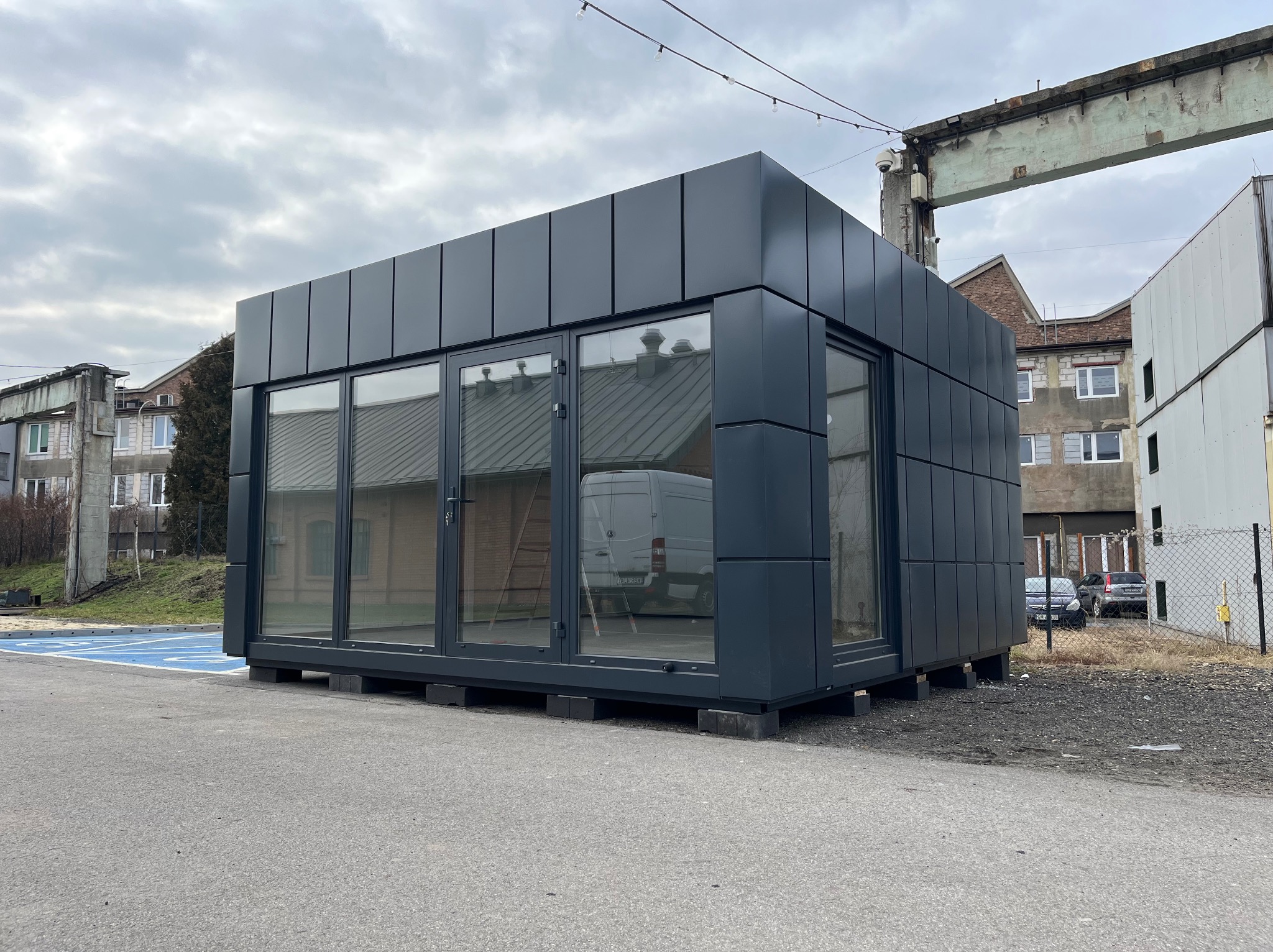 Nowoczesny pawilon handlowy z płyt warstwowych w kolorze grafitowym, z dużymi przeszkleniami, umieszczony na betonowych blokach, na tle przemysłowego krajobrazu z widocznym dźwigiem i budynkami.