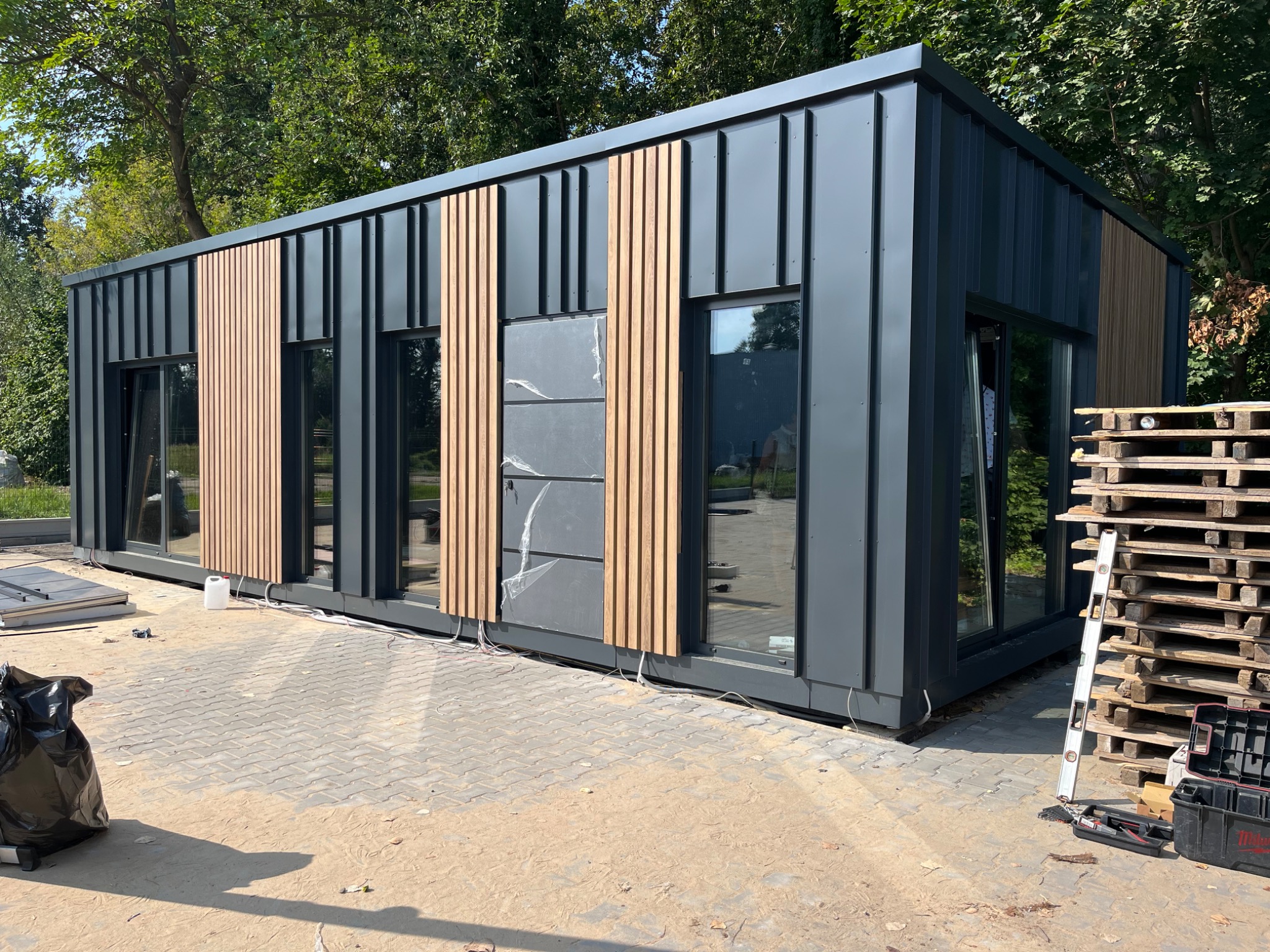 Nowoczesny, modułowy budynek biurowy z szarej blachy trapezowej i drewnianymi panelami dekoracyjnymi, częściowo jeszcze w folii ochronnej, widoczne palety i narzędzia budowlane sugerują etap montażu.