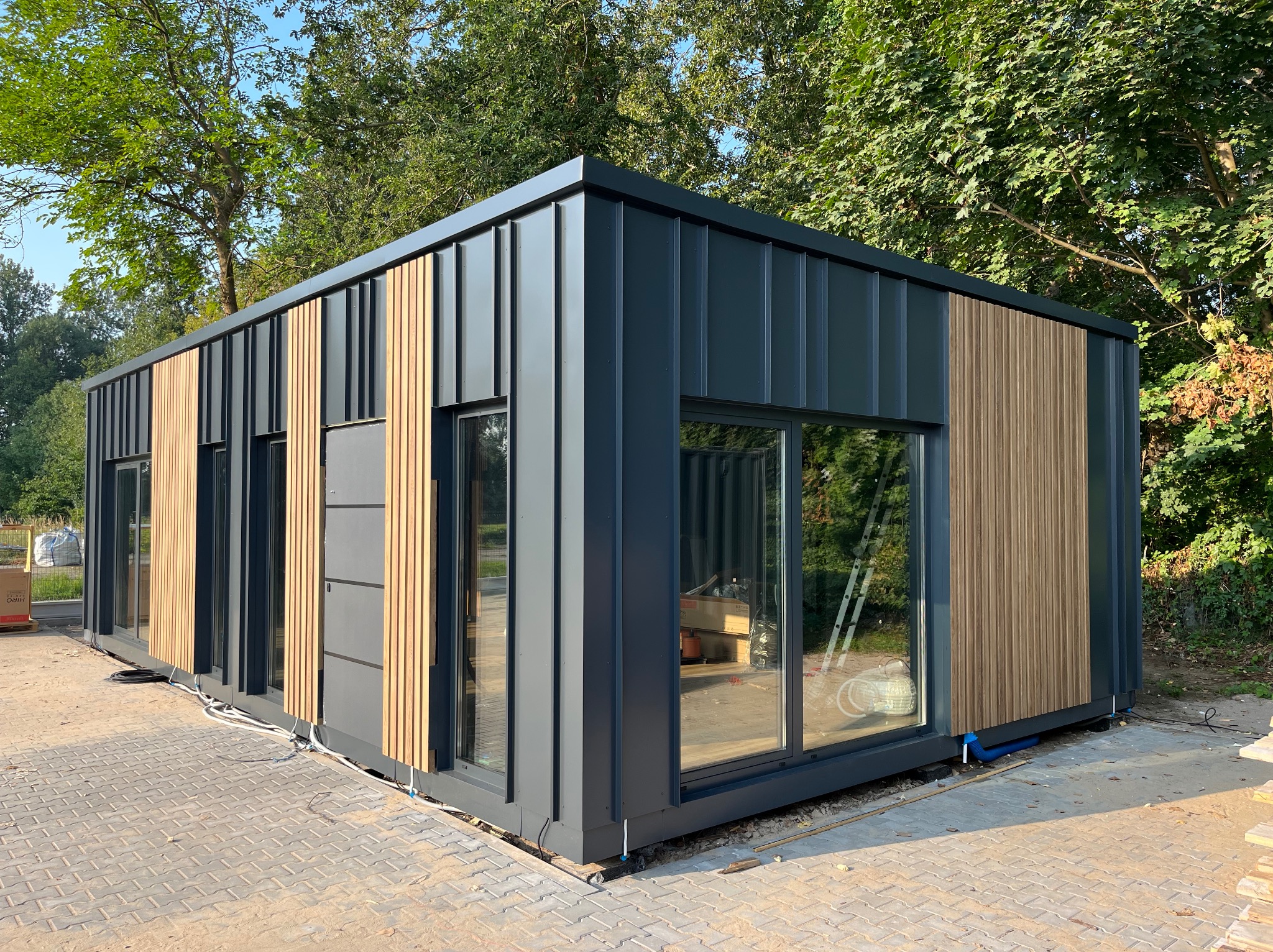 Gotowy modułowy budynek z płyt warstwowych w Białobrzegach, z elementami dekoracyjnymi z drewna, widoczne okna i drzwi, budynek posadowiony na kostce brukowej, otoczony zielenią.