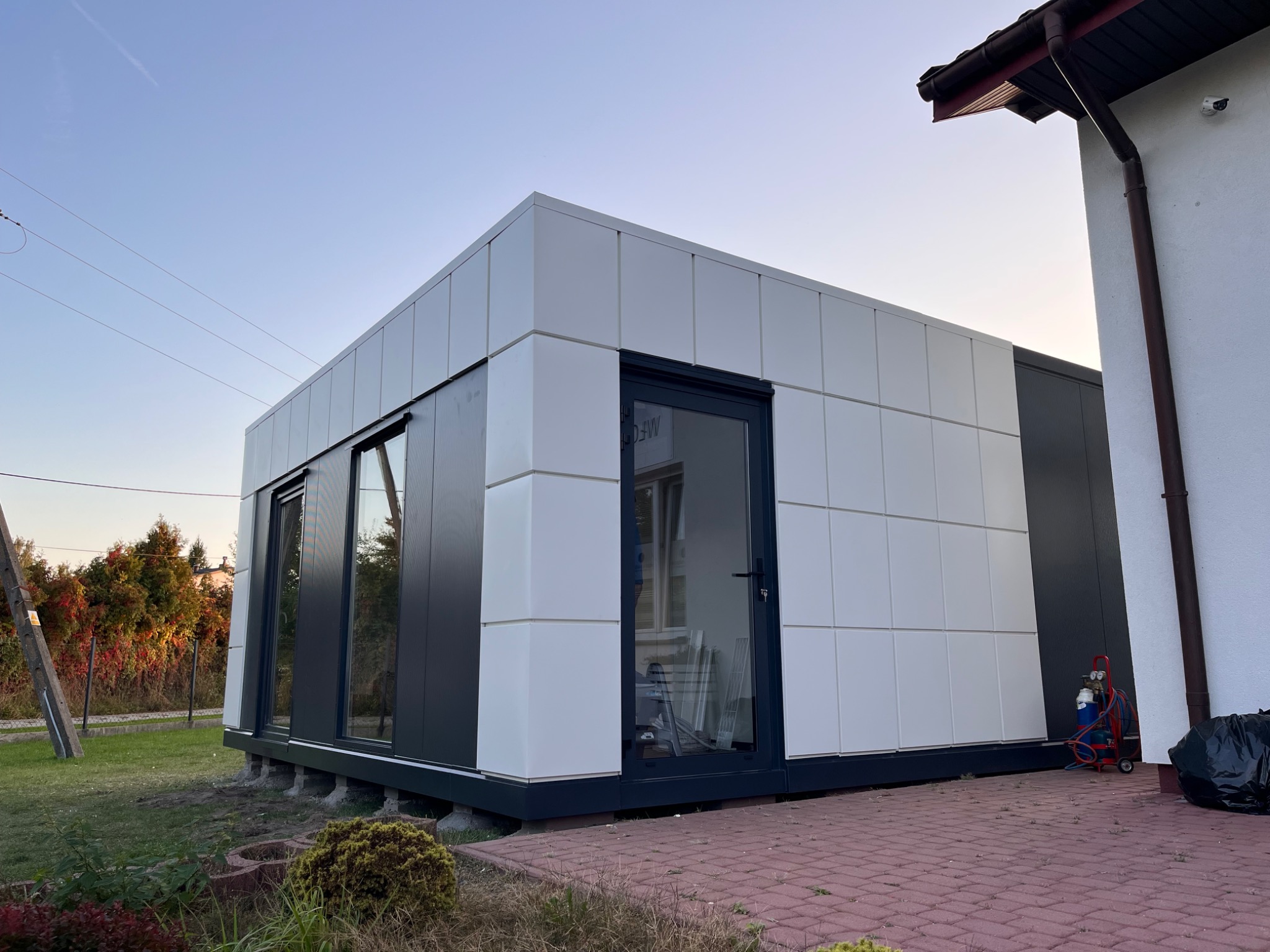 Gotowy modułowy budynek z białych i grafitowych płyt warstwowych, widoczne okna i drzwi z ciemną ramą, na zewnątrz butla z gazem i śmieci