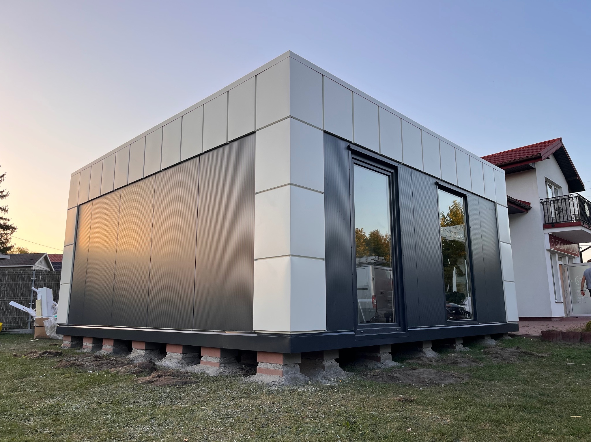 Nowoczesny, modułowy budynek z płyt warstwowych w trakcie montażu, widoczne ceglane podpory fundamentowe, szare panele ścienne i białe narożniki, tło: dom jednorodzinny i zachodzące słońce.