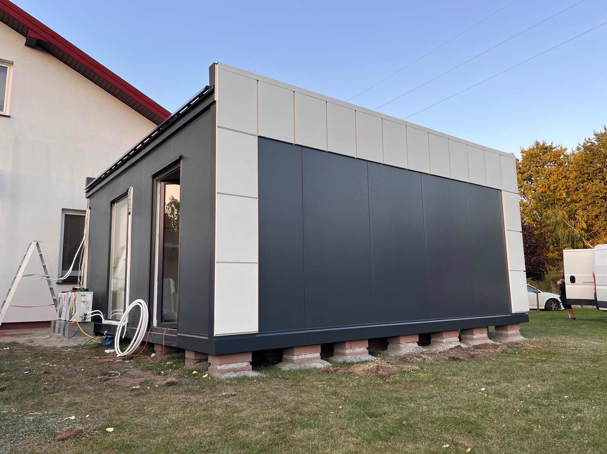 Montaż modułowego budynku z płyt warstwowych na bloczkach betonowych, widoczne instalacje klimatyzacji, częściowo otwarte drzwi wejściowe, budynek sąsiaduje z domem jednorodzinnym.