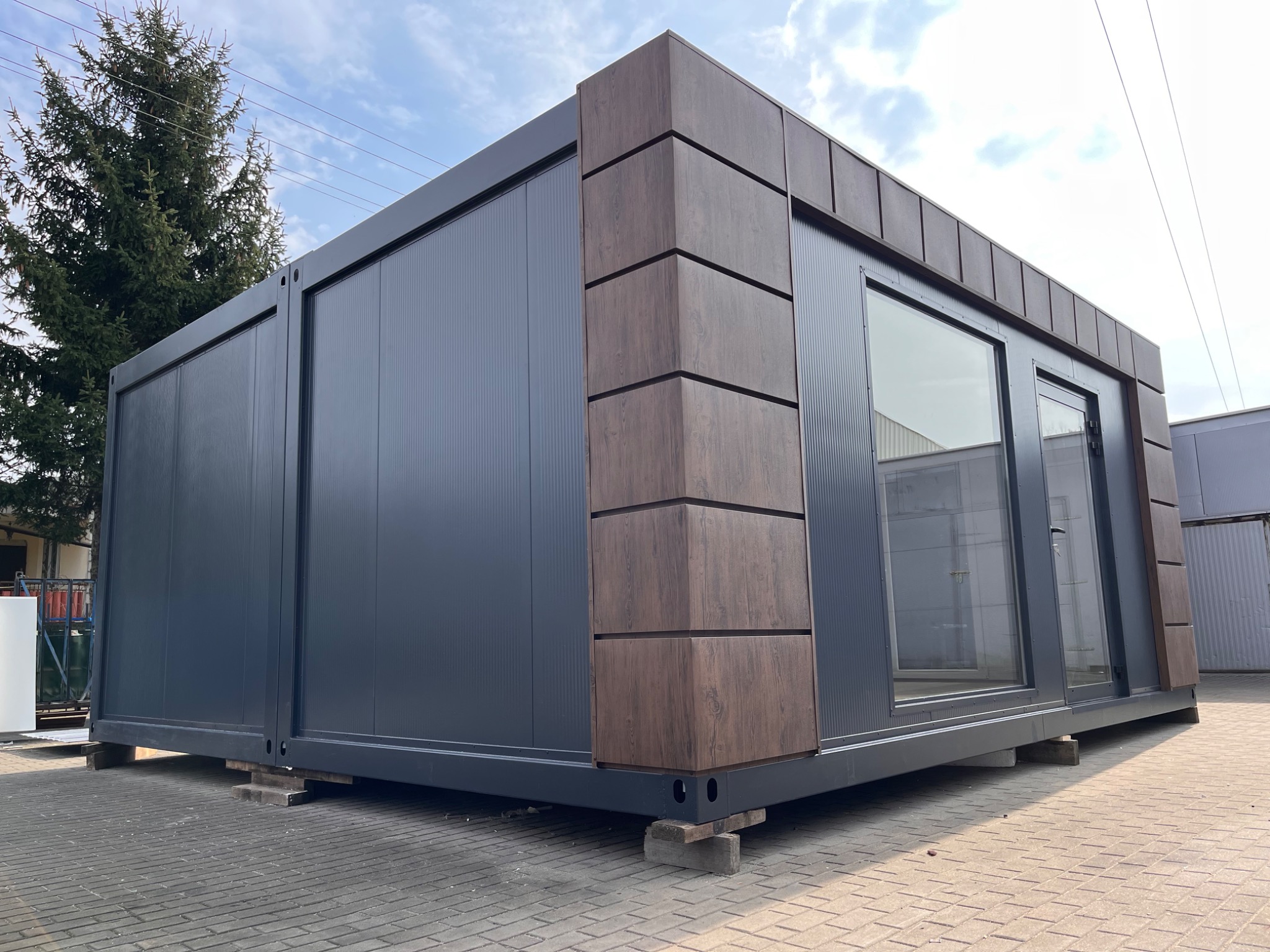 Nowoczesny modułowy budynek z szarymi ścianami z płyty warstwowej i dekoracyjnymi panelami imitującymi drewno, ustawiony na bloczkach betonowych na kostce brukowej.