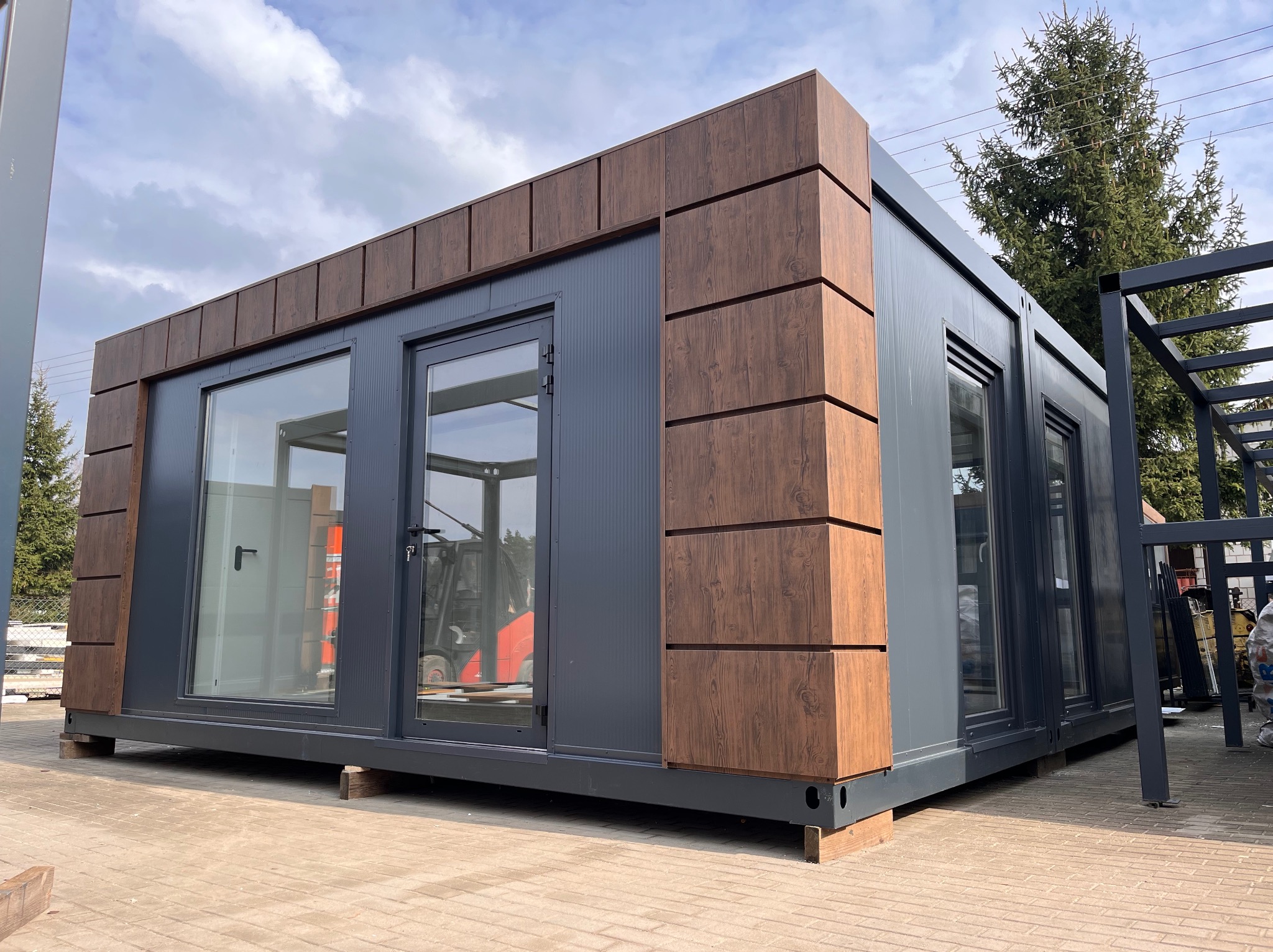 Gotowy modułowy budynek z płyt warstwowych, wykończony panelami imitującymi drewno, umieszczony na kostce brukowej, z widocznym w tle wózkiem widłowym.