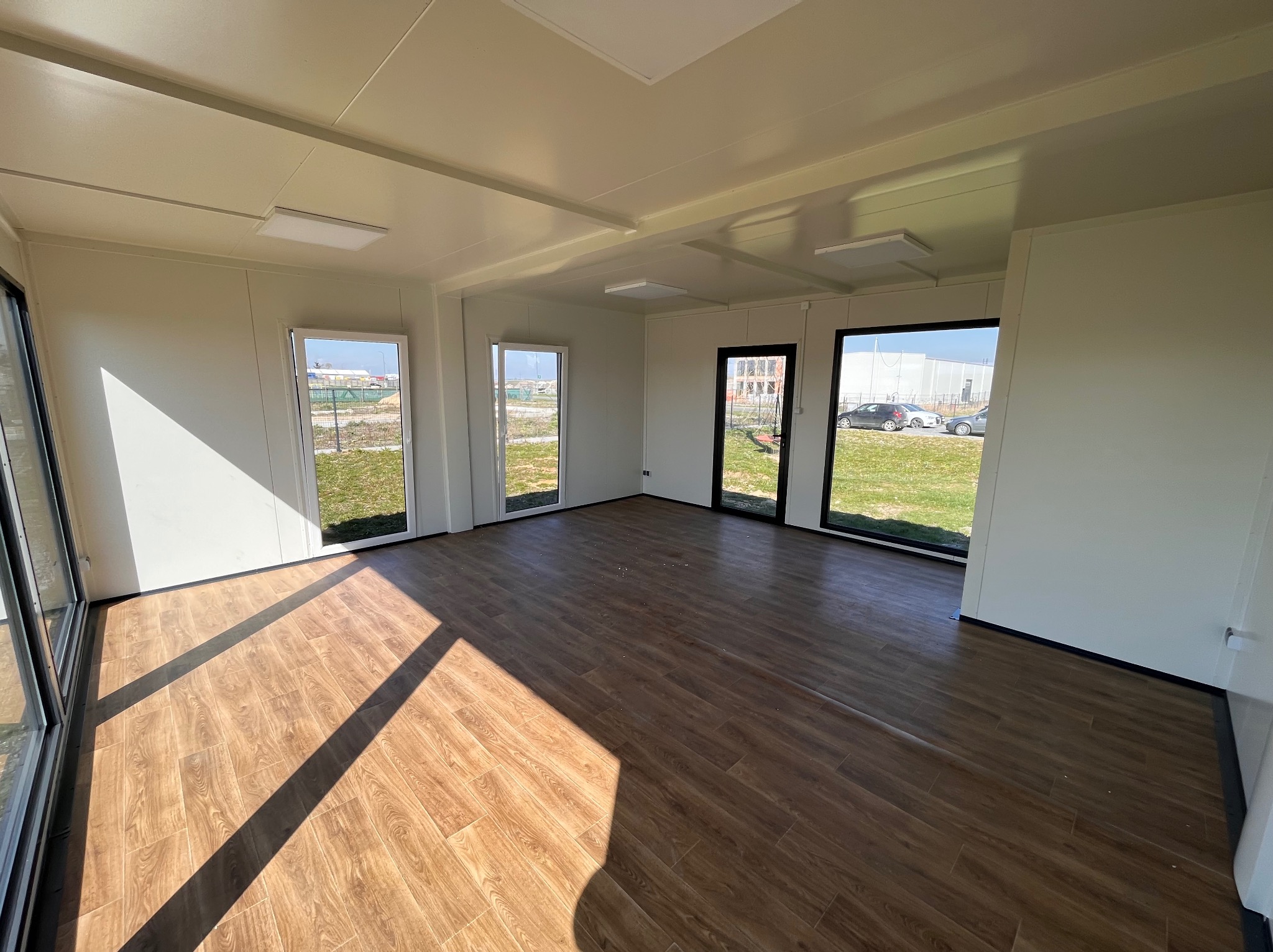 Wnętrze modułowego biura z panelami ściennymi, podłogą imitującą drewno i dużymi oknami w czarnych ramach, wpadające promienie słoneczne.