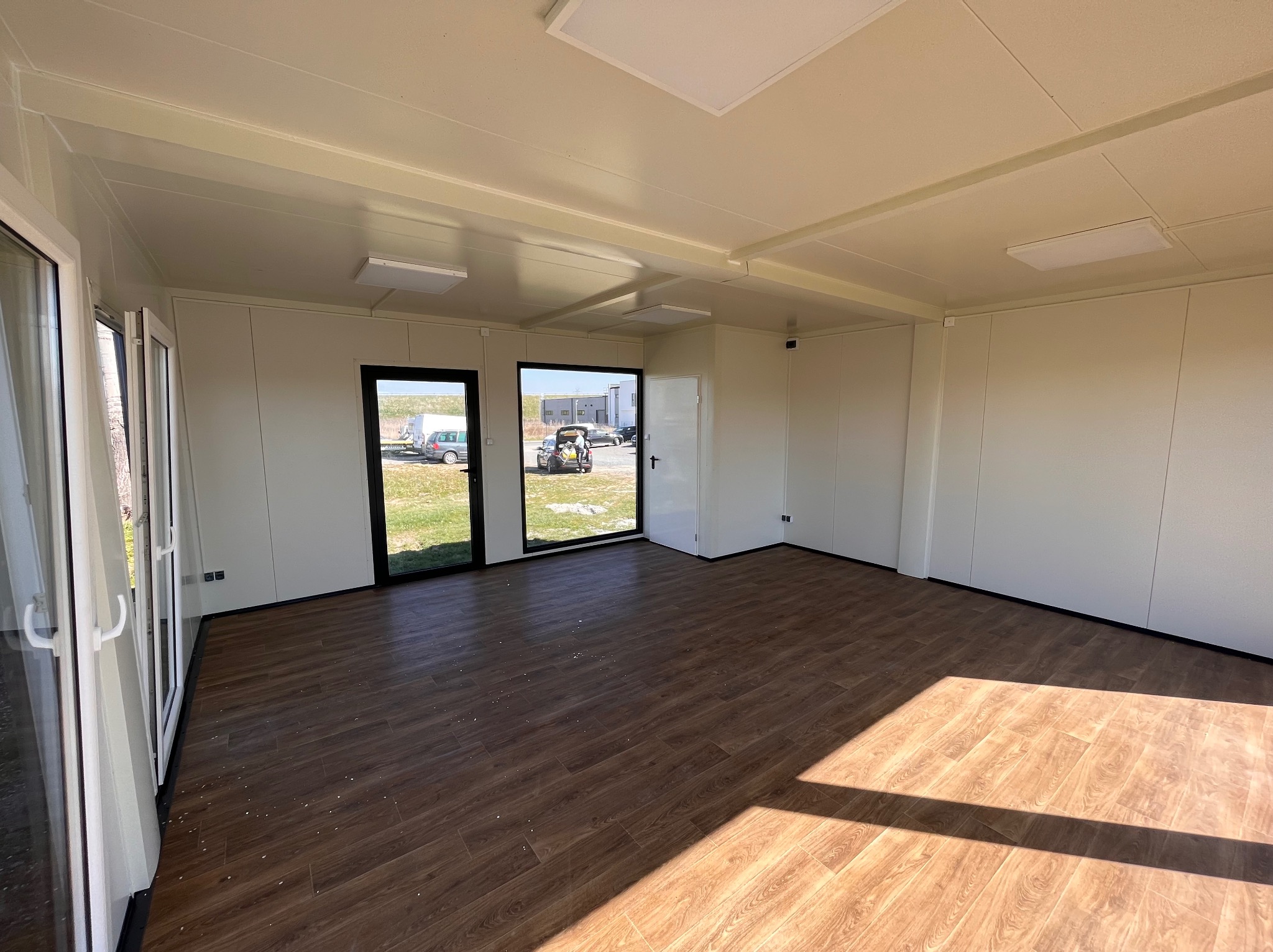 Wnętrze modułowego biura z panelami ściennymi, oknami i drzwiami z widokiem na zewnątrz, podłoga wyłożona panelami imitującymi drewno, oświetlenie ledowe w suficie.