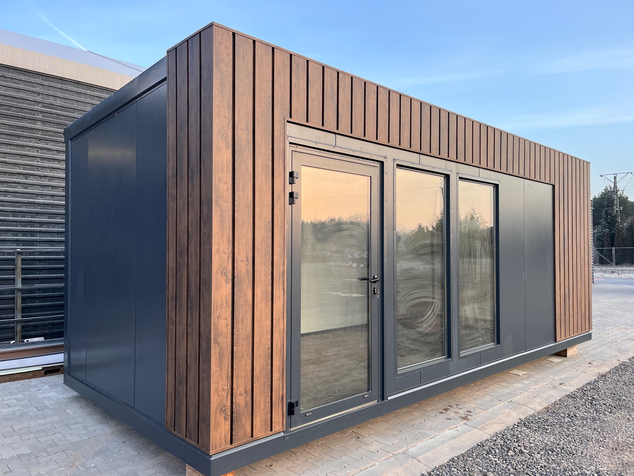 Nowoczesny pawilon biurowy z płyt warstwowych w kolorze grafitowym i okładziną drewnopodobną, widok z zewnątrz na tle błękitnego nieba.