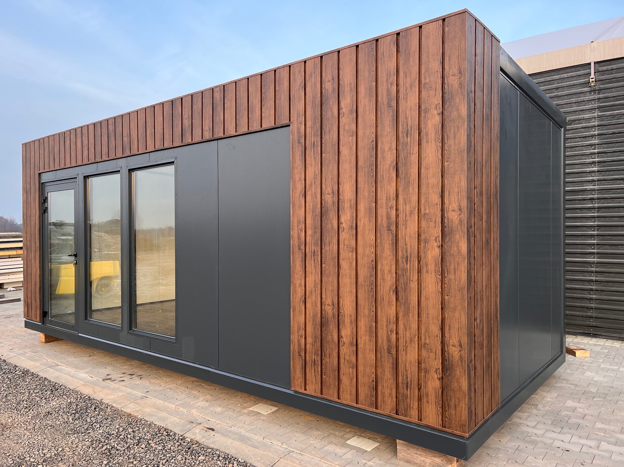 Nowoczesny kontener biurowy z panelami imitującymi drewno i szarymi panelami, ustawiony na kostce brukowej, widoczne okna i drzwi.