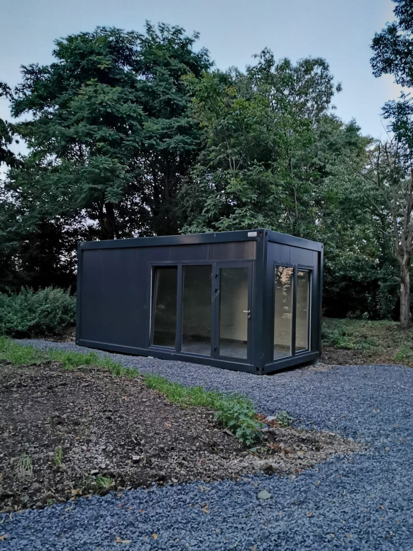 Nowoczesny, ciemnoszary pawilon modułowy z dużymi oknami, ustawiony na żwirowym podłożu, otoczony zielenią drzew