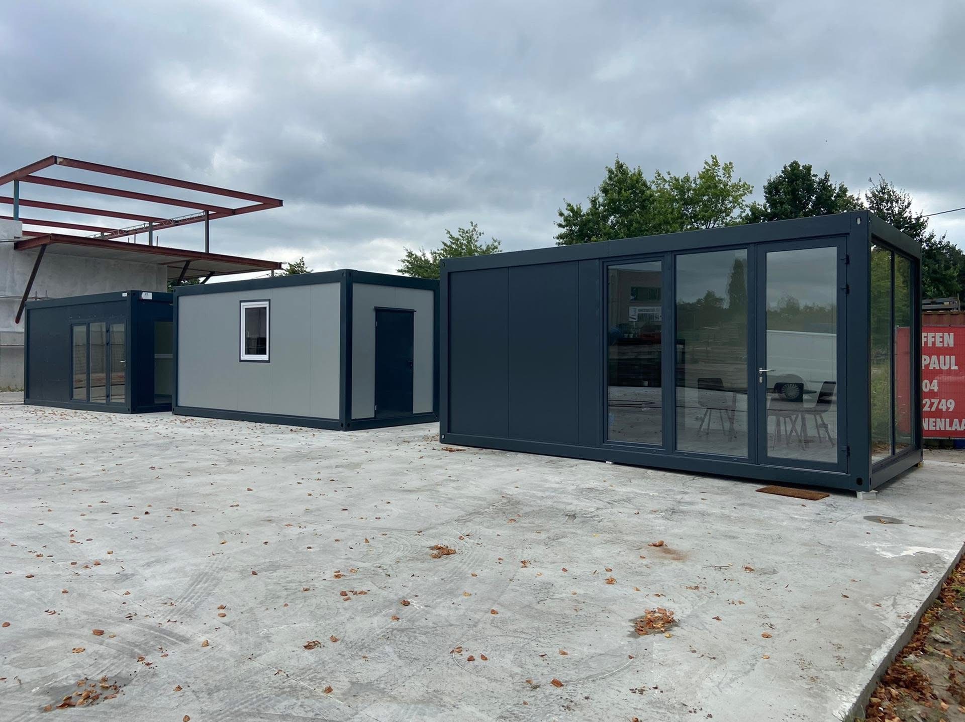 Trzy modułowe kontenery biurowe w różnych kolorach (szary, grafitowy) z oknami i drzwiami, ustawione na betonowym placu pod pochmurnym niebem.