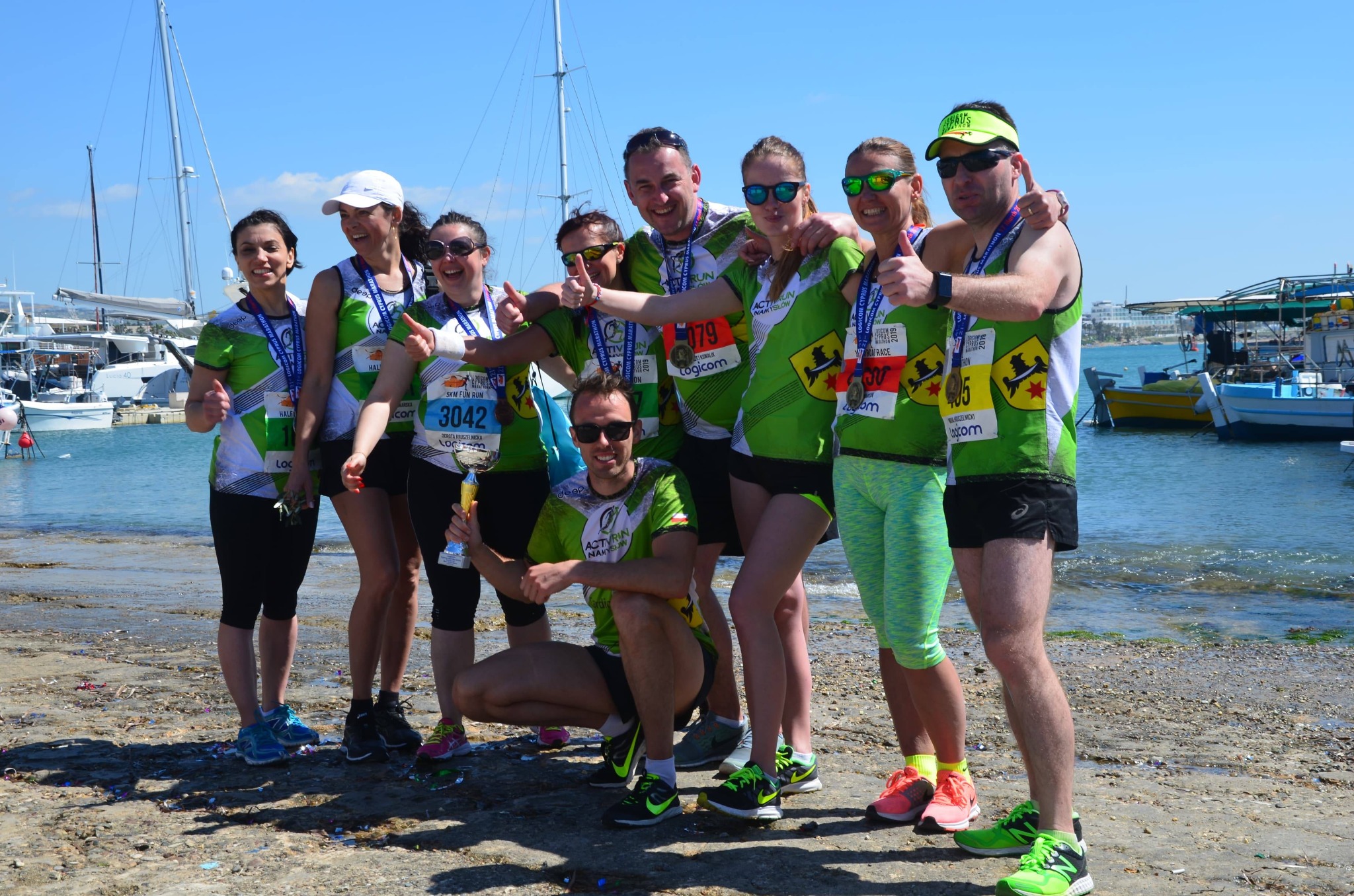 Grupa uśmiechniętych biegaczy z Namysłowa świętuje zwycięstwo w biegu, prezentując medale i puchar na tle portu jachtowego.