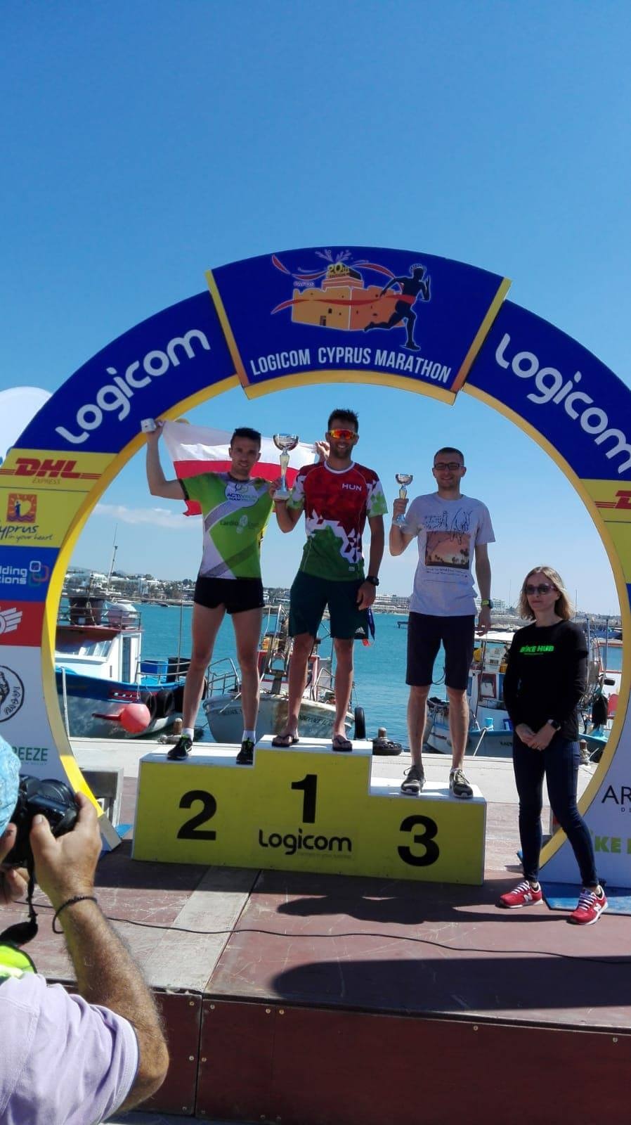 Trzej mężczyźni na podium z trofeami, po ukończonym maratonie Logicom Cyprus Marathon, w tle port z łodziami i błękitne niebo.