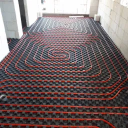 Czerwone rury ułożone w spiralny wzór na czarnej macie podłogowej systemu ogrzewania w surowym wnętrzu budynku w trakcie budowy, widoczna taśma klejąca.