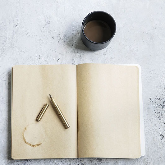 Otwarty notatnik z czystymi stronami, pióro wieczne i ślad po kawie, obok czarny kubek z kawą, na szarym betonowym tle.