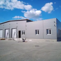 Nowoczesny budynek przemysłowy z szarej blachy panelowej, widoczne dwie drabiny opierające się o ściany, błękitne niebo z białymi chmurami w tle.