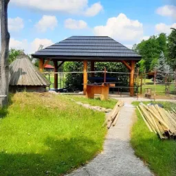 Altana z ciemnym, nowym dachem z blachy na tle błękitnego nieba z chmurami, widoczne deski i przygotowany teren.