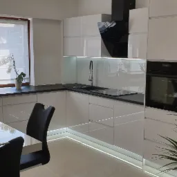 Nowoczesna, jasna kuchnia z białymi lakierowanymi frontami, czarnym blatem, oświetleniem LED pod szafkami i sprzętem AGD w zabudowie, widok na stół z krzesłami.