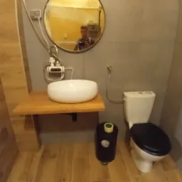 Ukończona łazienka z umywalką nablatową na drewnianym blacie, toaleta z czarną deską, lustro okrągłe z odbiciem osoby robiącej zdjęcie, płytki imitujące drewno na podłodze i ścianie, szare płytki...