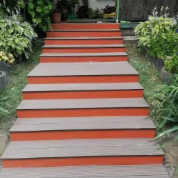 Schody zewnętrzne z desek kompozytowych, pomalowane na pomarańczowo, prowadzące do domu z zielenią w tle.