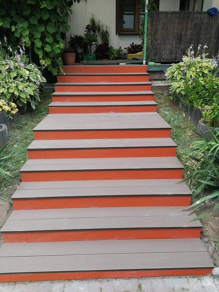 Schody zewnętrzne z desek kompozytowych, pomalowane na pomarańczowo, prowadzące do domu z zielenią w tle.