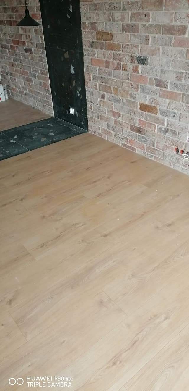 Jasne panele podłogowe imitujące drewno ułożone w pomieszczeniu z ceglaną ścianą i czarnym wykończeniem przy wejściu.