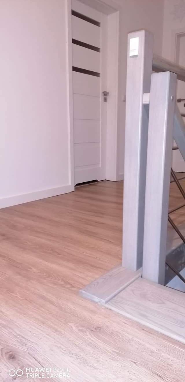 Jasne panele podłogowe ułożone wewnątrz pomieszczenia, widoczna biała ściana, drzwi i fragment schodów z białą balustradą.