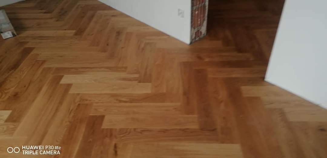 Ułożony parkiet w jodełkę na podłodze w jasnym odcieniu drewna, widoczne fragmenty białej ściany i niezabezpieczony narożnik ściany.