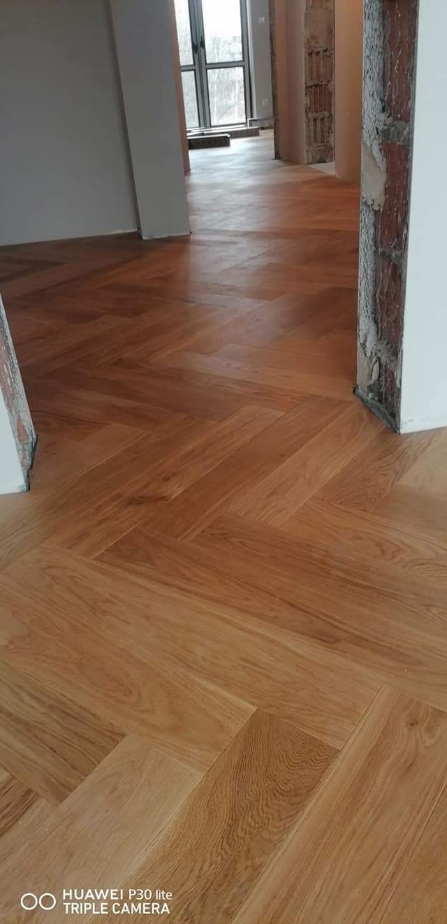 Ułożony parkiet w jodełkę, z widocznymi surowymi fragmentami ścian i kolumn, jasne drewno, naturalne oświetlenie wpadające przez okno w tle.