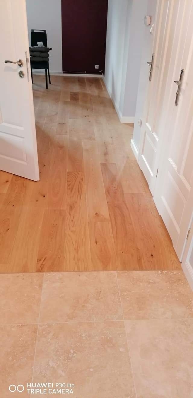 Jasne, drewniane panele podłogowe przechodzące w beżowe płytki ceramiczne, widoczne białe drzwi i fragment pomieszczenia z krzesłem w tle.