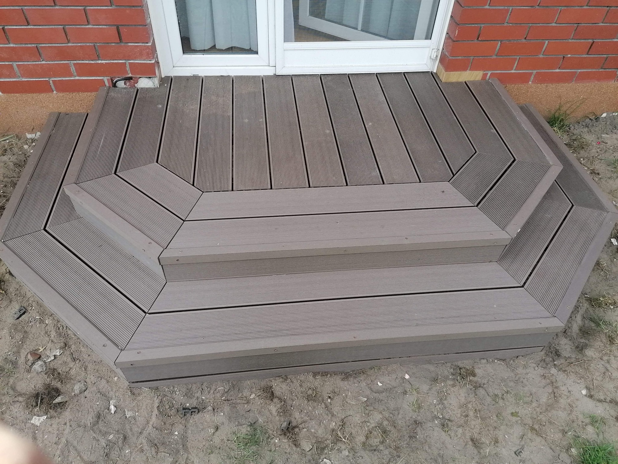 Schody wejściowe wykonane z kompozytowych desek tarasowych, prowadzące do drzwi balkonowych, z widoczną ceglaną elewacją budynku w tle.